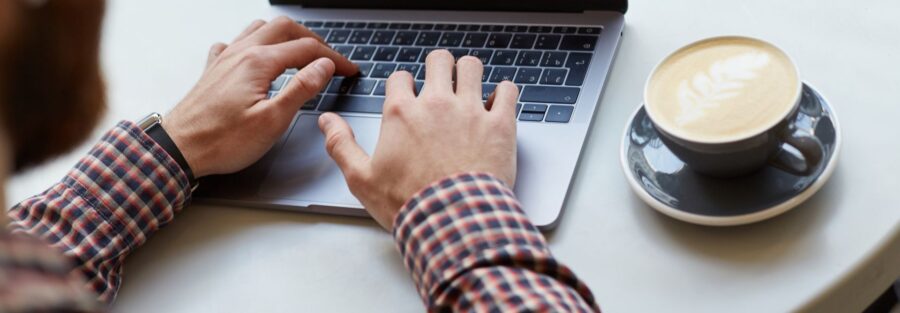 In A Cafe On A White Table, Men's Hands Are Working On The Keyboard Of The Laptop